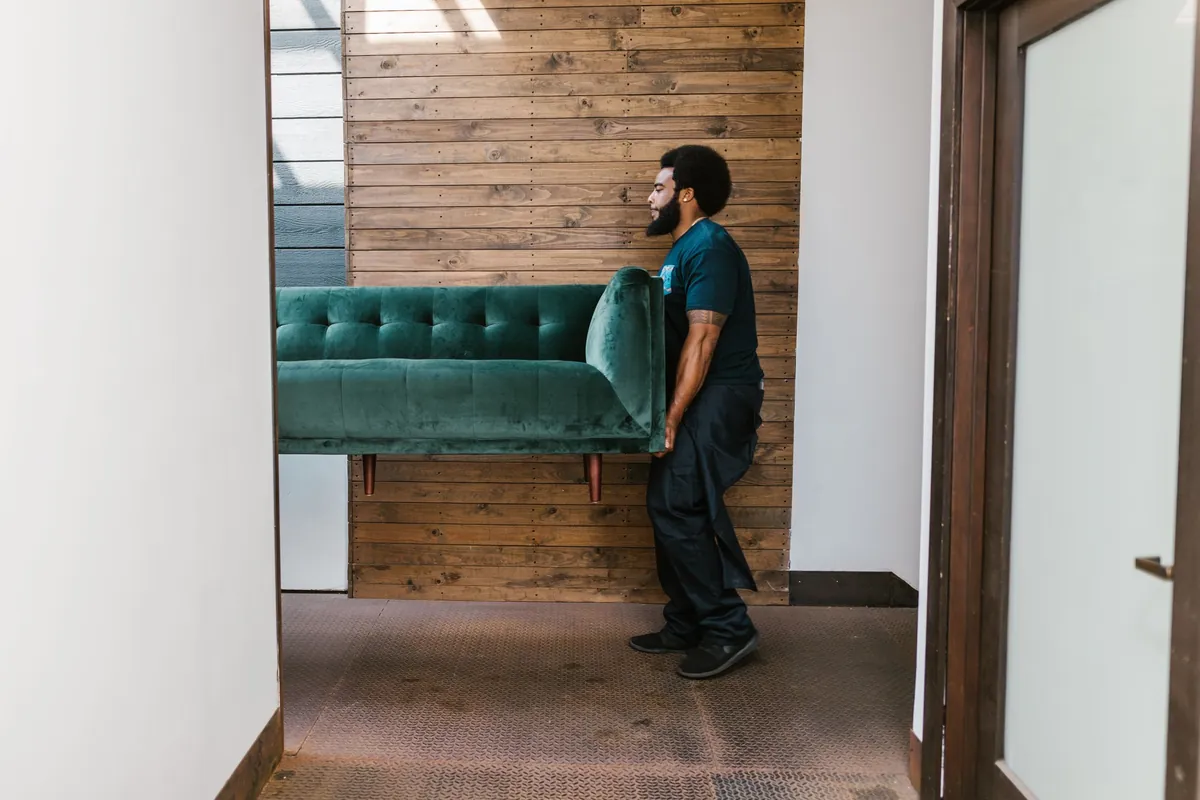 A crew member carrying a single green sofa out through a hallway